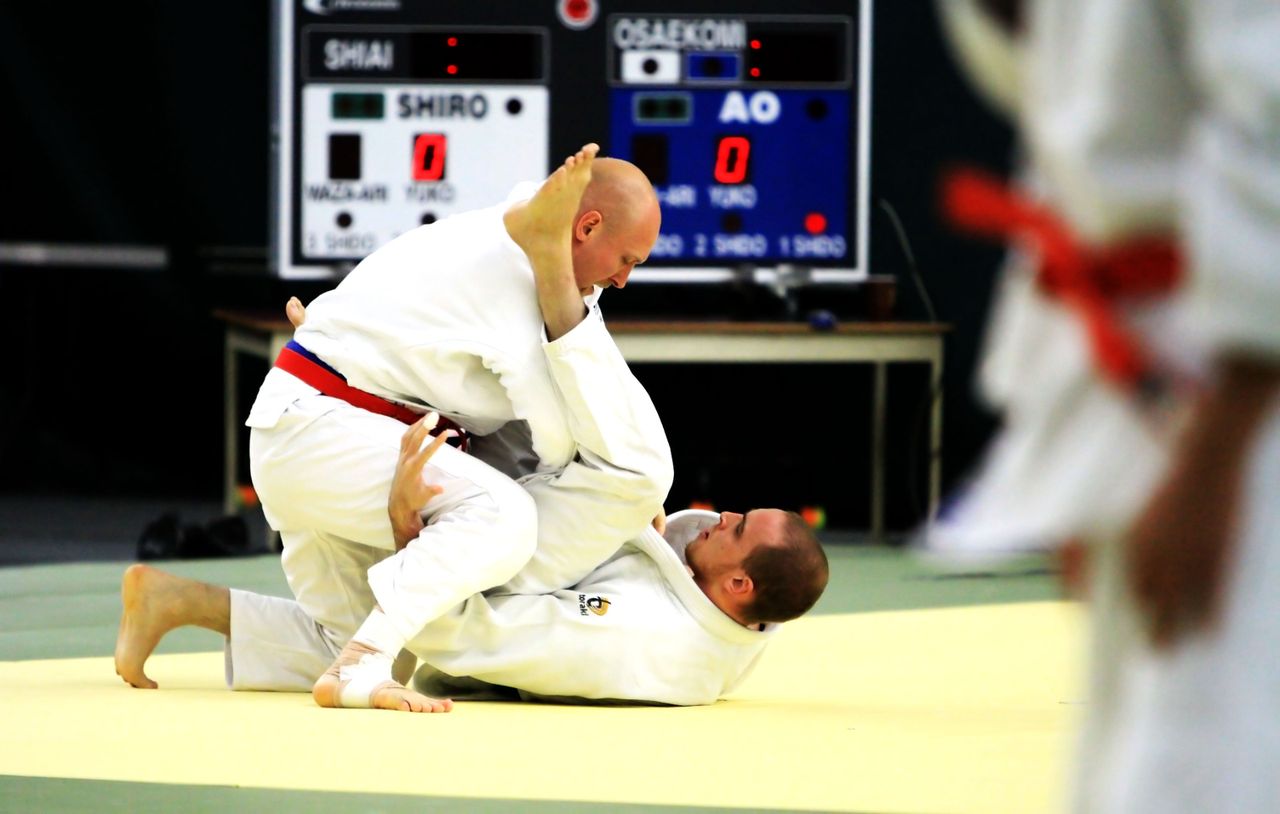 Two adult judoka in groundwork beneath a competition scoreboard